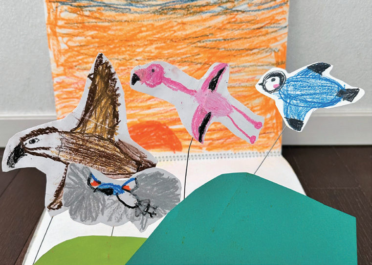 子どもが描いて切り抜いた鳥たちが、夕焼けの空を背景に飛んでいる立体作品